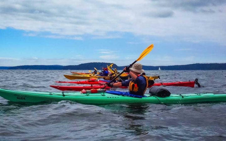 a line of colorful kayaks are paddled by outward bound students on a calm body of water. There appear to be mountains in the background.